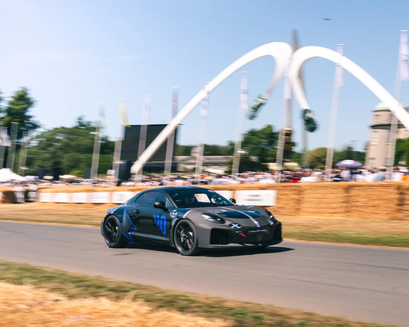 Matte black Alpine race car with blue number 70 and star decal driving on a track with hay bales and crowd in background.