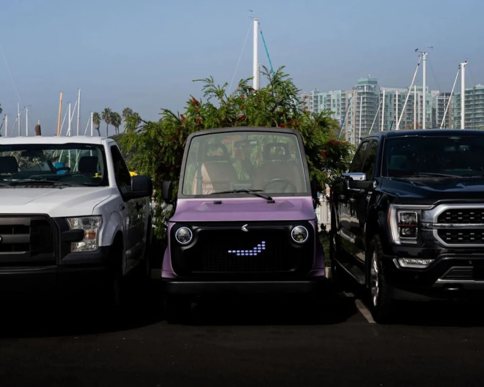 Small purple electric car with a digital smiley face on the grille parked between two large pickup trucks.
